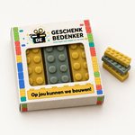 Vierkant bedrukt doosje met rechthoekig venster en drie chocolade bouwstenen in lego-stijl, met gestapelde bouwstenen ernaast
